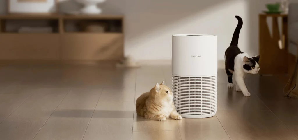 A cylindrical white Xiaomi smart air purifier standing on a wooden floor in a living room, with a fluffy ginger cat lying beside it and a black-and-white cat walking past it in the background.
