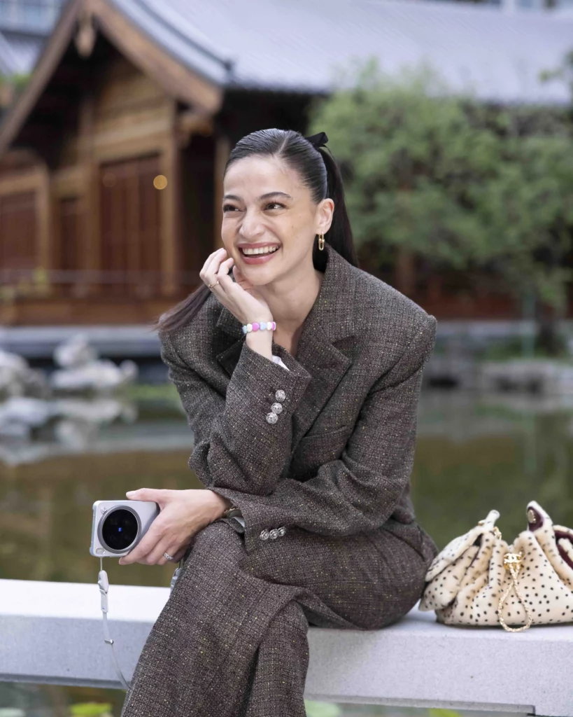 Anne Curtis holding a gray vivo smartphone fitted with a telephoto extender lens while sitting on a ledge outdoors in front of a traditional Chinese-style building, smiling with her hand resting on her cheek.