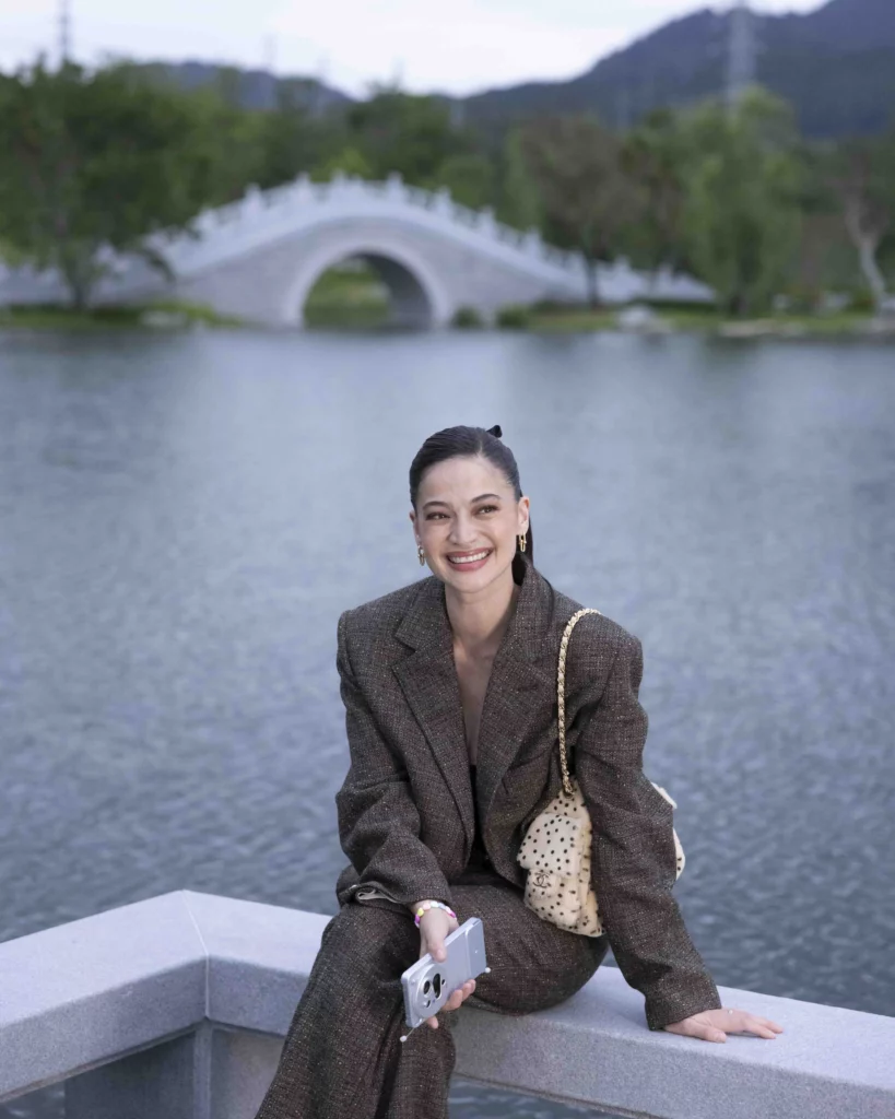 Anne Curtis smiling while sitting on a stone wall in front of a scenic pond and a traditional white stone bridge in the background at the vivo Global HQ campus.