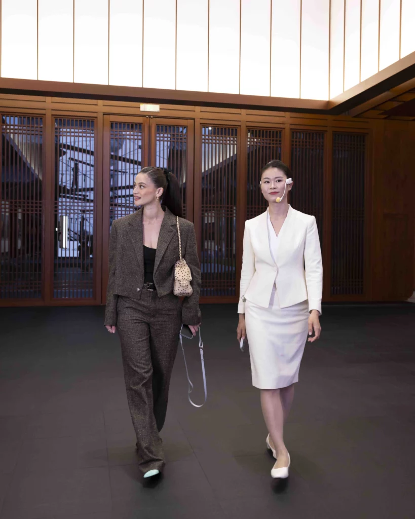 Anne Curtis walking through a modern wooden interior hallway alongside a tour guide wearing a white skirt suit and a headset microphone at the vivo headquarters.