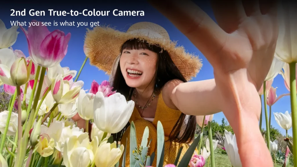 A smiling woman wearing a straw hat posing in a field of vibrant pink, white, and yellow tulips while taking a low-angle selfie. Text overlay reads: "2nd Gen True-to-Colour Camera. What you see is what you get."