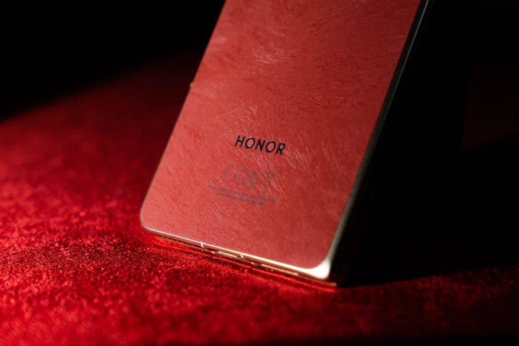 Close-up view of the lower back panel of a red textured HONOR Magic V6 smartphone with a gold metallic edge resting on a red surface, displaying the centered black HONOR logo.