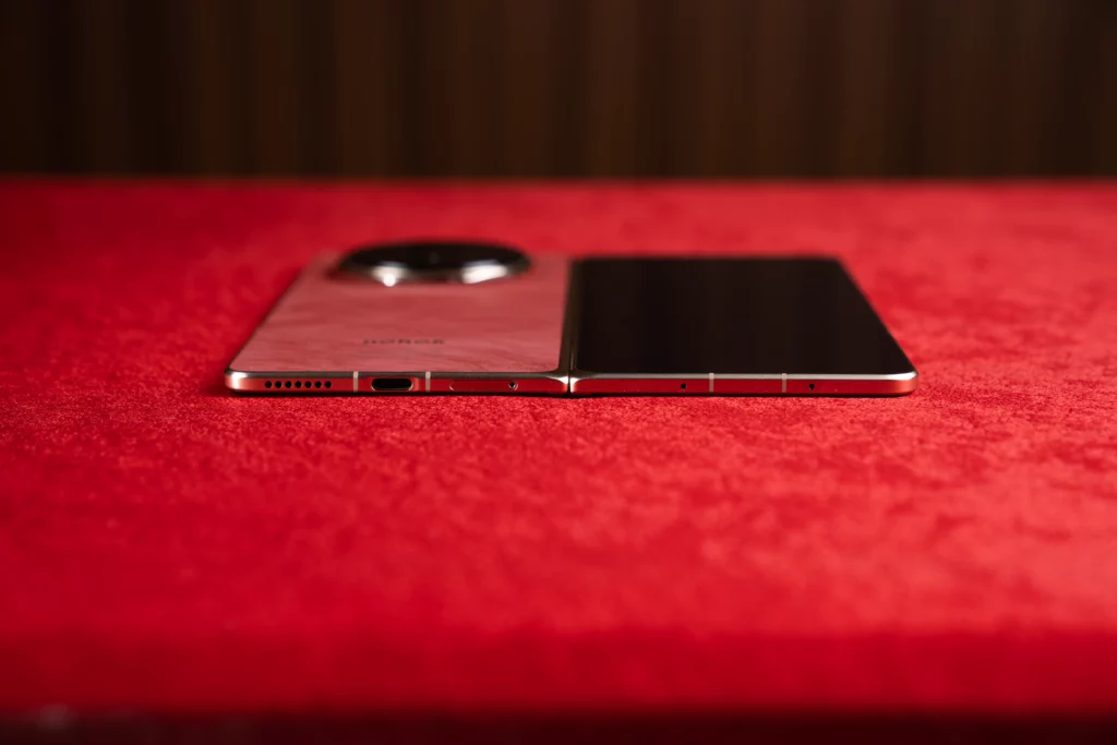 Bottom edge view of the unfolded HONOR Magic V6 lying flat on a red surface, showing the USB Type-C charging port, speaker grille, and microphone cutout.