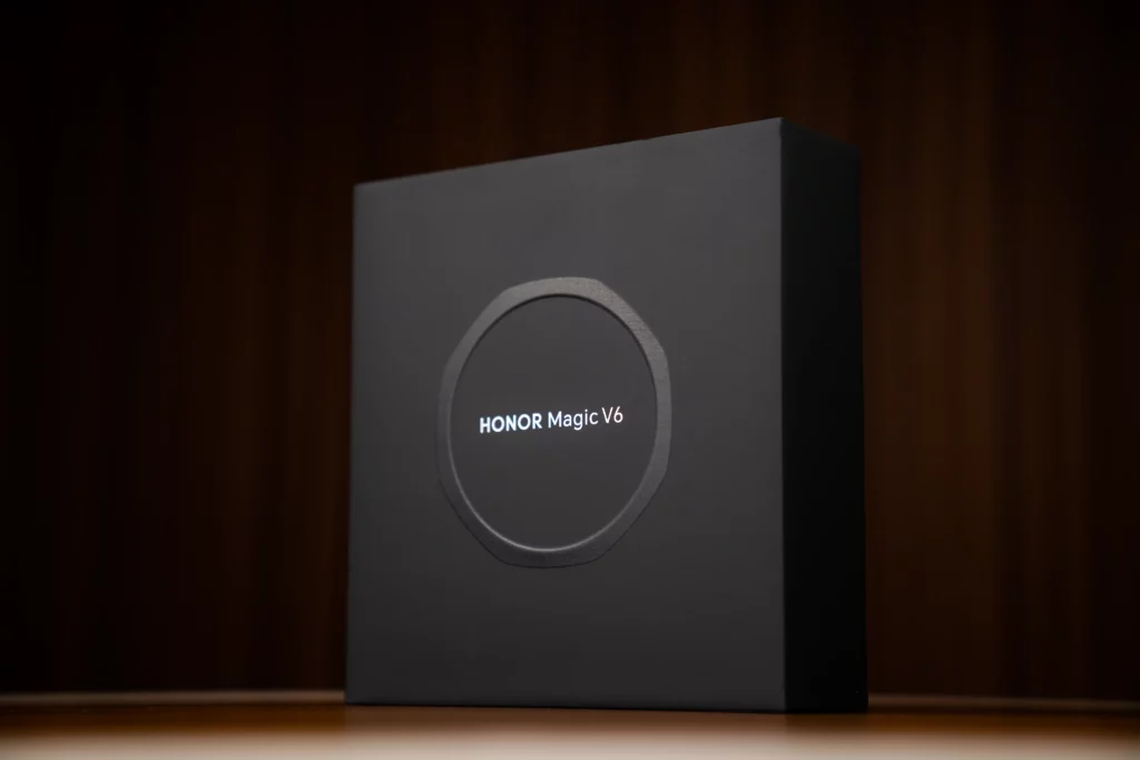 Matte black square retail box for the HONOR Magic V6 smartphone standing upright on a wooden surface, featuring a circular ring design in the center with the model name printed inside.