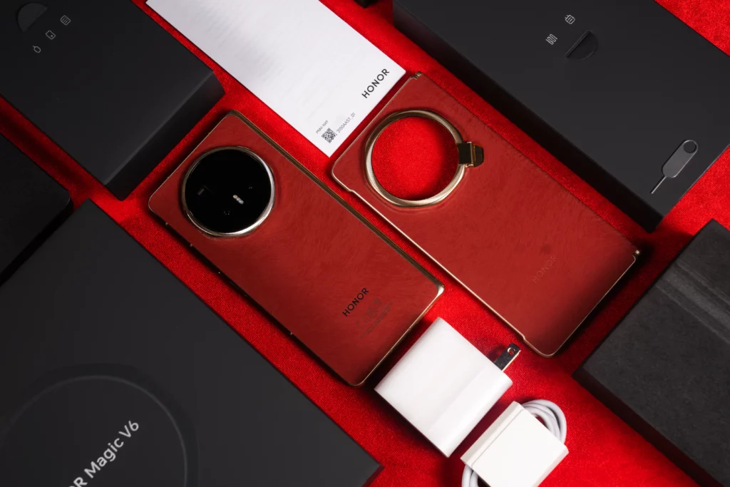 Flat-lay layout of the red HONOR Magic V6 foldable smartphone and its unboxing accessories spread across a red felt background, including a matching red phone case with a integrated ring stand, white charging brick, charging cable, black box insert, and SIM ejector tool.