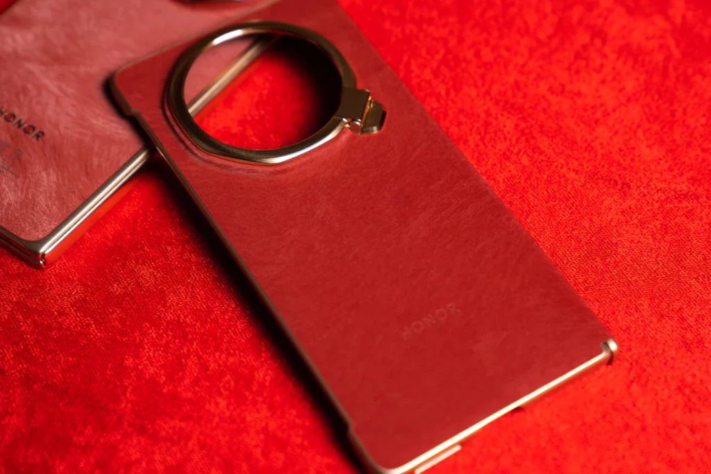 Close-up of a red textured phone case resting on a red surface, featuring a raised gold circular camera module cutout with a fold-out ring stand mechanism.