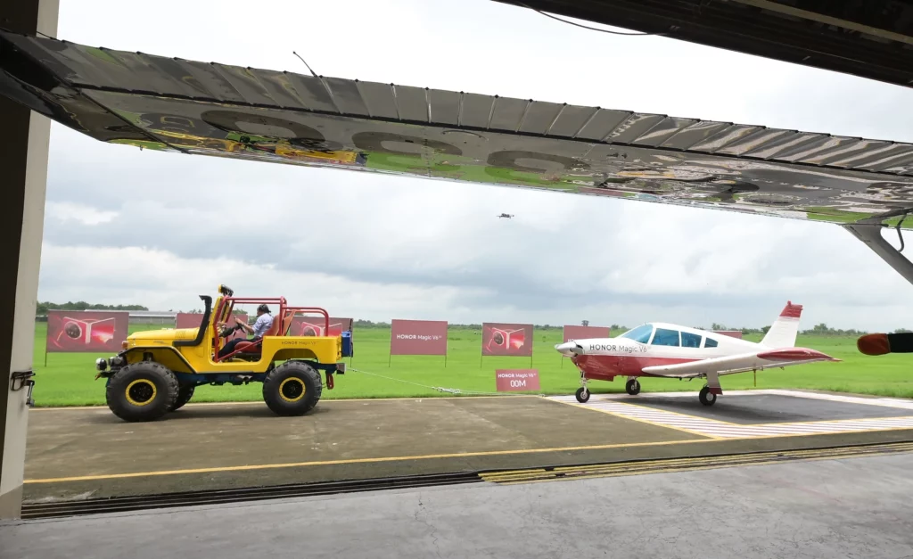 Wide shot taken from inside an airfield hangar showing a yellow vehicle towing a light aircraft on the tarmac under a overcast sky.