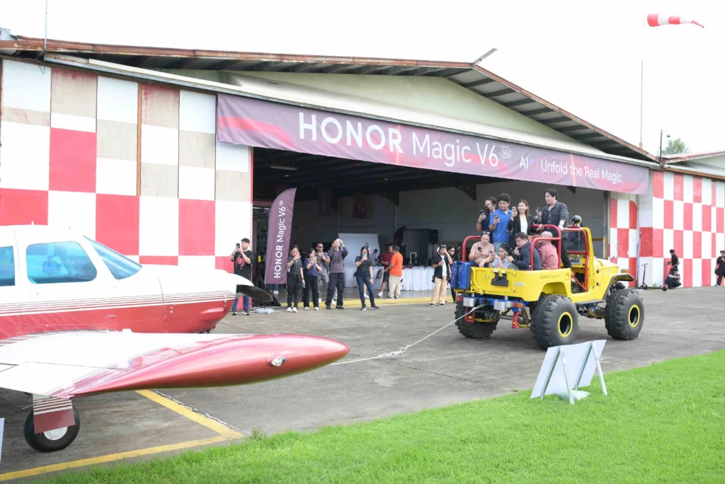 Wide view of a yellow vehicle filled with onlookers pulling a red and white light aircraft outside an airplane hangar decorated with HONOR Magic V6 banners.