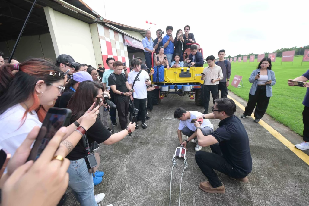 A crowd of spectators and media filming two men securing an HONOR Magic V6 foldable smartphone into a metal towing rig attached to steel cables on an airfield tarmac.
