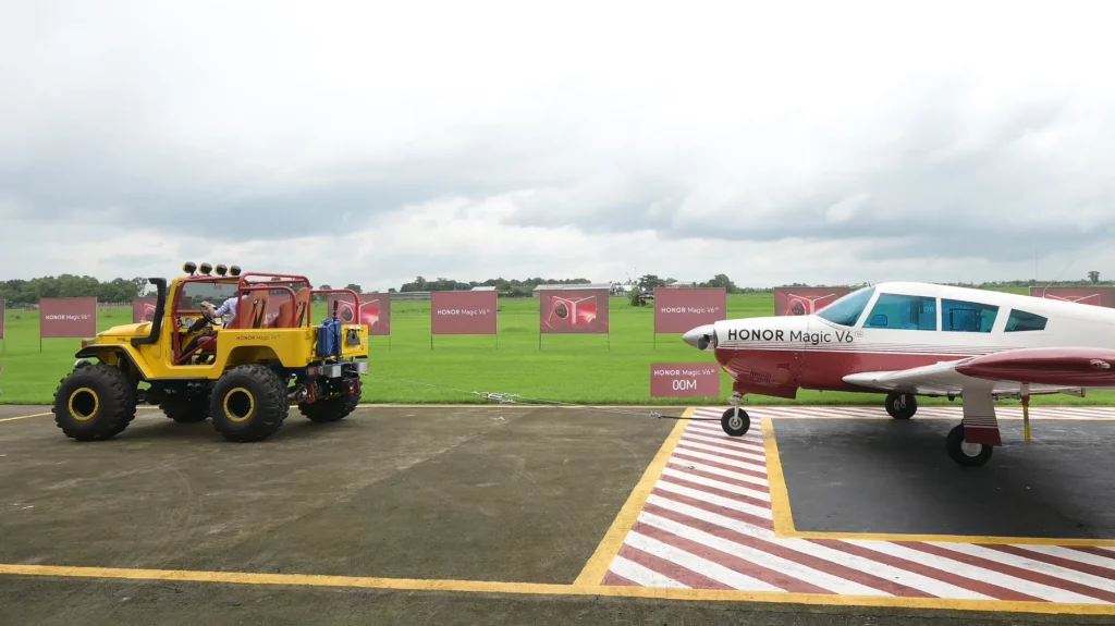 Side profile of a yellow off-road vehicle connected via tow cable and an HONOR Magic V6 smartphone to a red and white light aircraft on a runway.