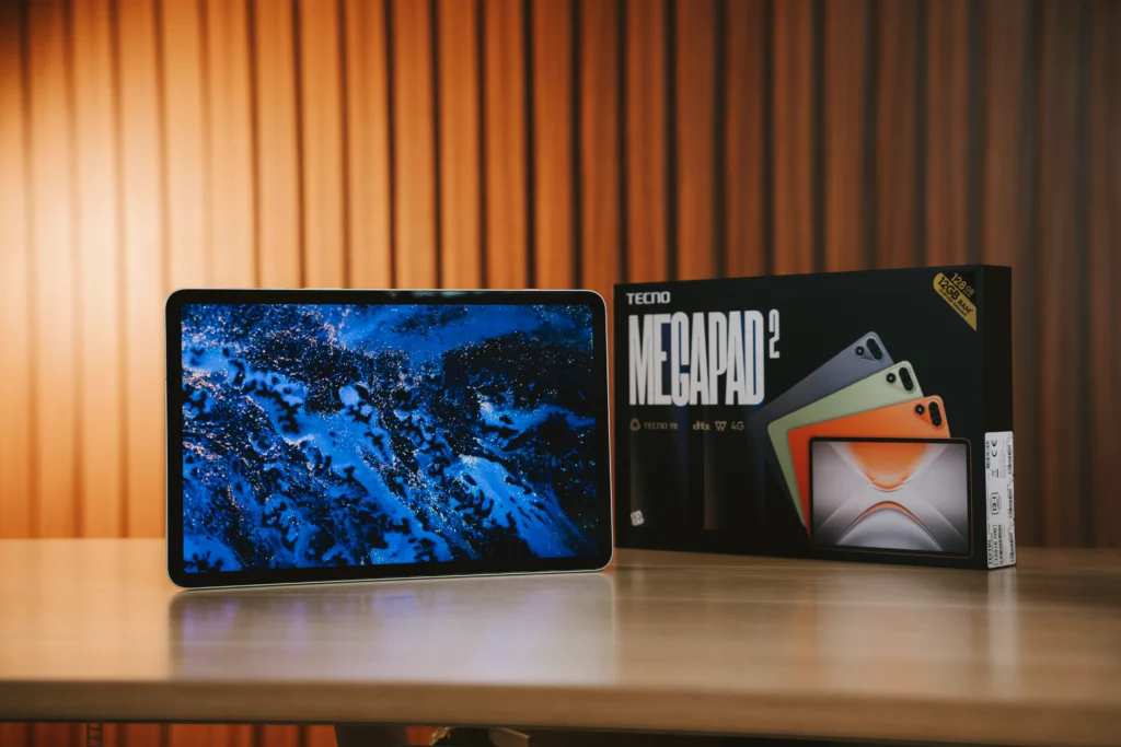 The TECNO MEGAPAD 2 tablet rests on a wooden table displaying a vibrant blue abstract wallpaper, placed beside its official retail box.
