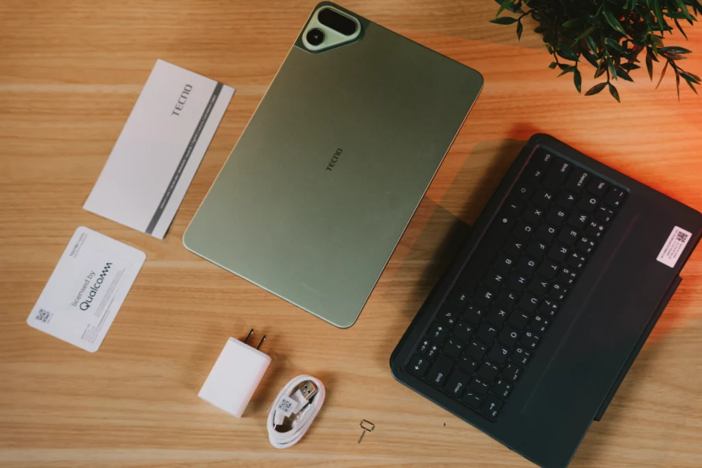 Flat lay photo of the light green TECNO MEGAPAD 2 surrounded by accessories, including a power adapter, USB cable, SIM ejector tool, warranty cards, and a black detachable keyboard case.
