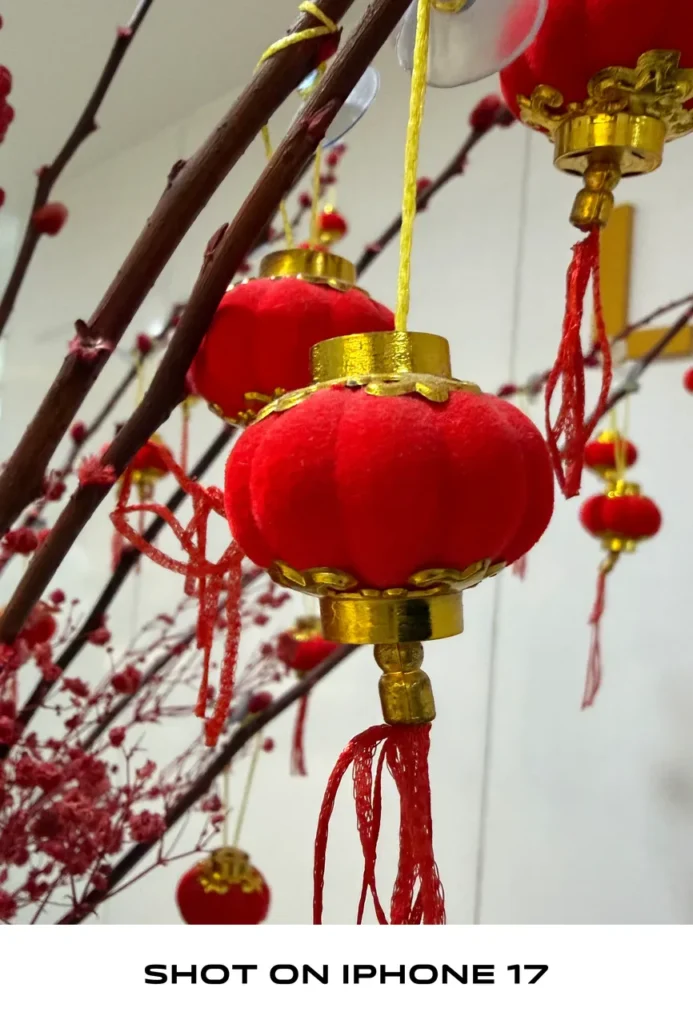 Close-up photo of small red mini Chinese lanterns with gold trim hanging on branches, labeled "SHOT ON IPHONE 17".