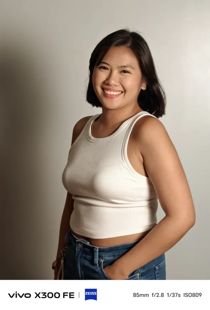Portrait photo of a woman in a white ribbed tank top smiling against a neutral wall, captured on the vivo X300 FE with ZEISS camera watermarks.