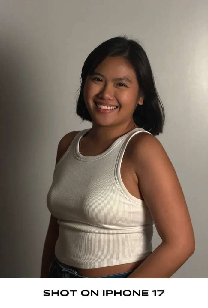 Portrait photo of a woman in a white ribbed tank top smiling against a neutral wall, labeled "SHOT ON IPHONE 17".