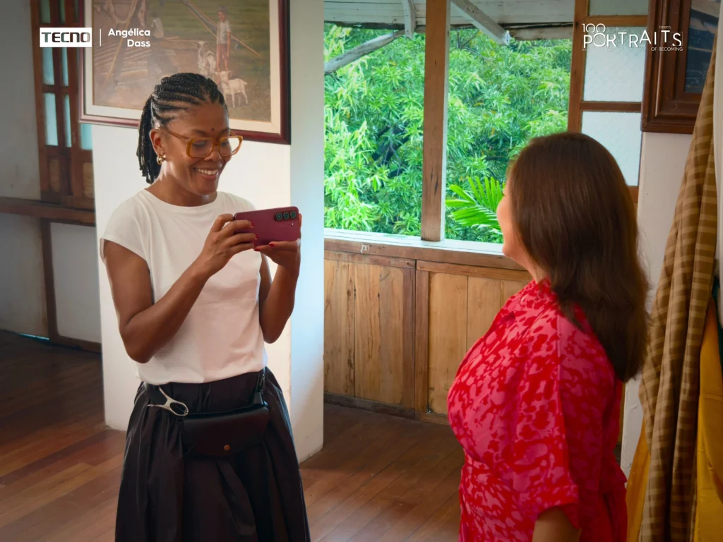 Close-up shot of Angélica Dass holding a pinkish-burgundy TECNO smartphone to photograph a woman wearing a red patterned dress, featuring logos for TECNO, Angélica Dass, and 100 Portraits of Becoming.