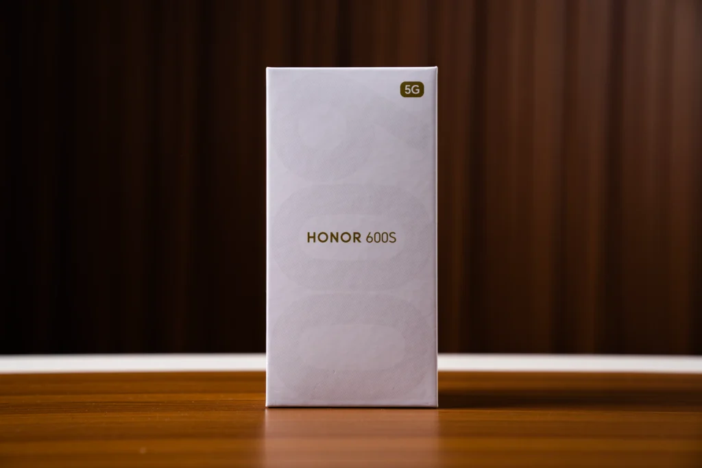 The front of the white retail packaging box for the HONOR 600S 5G resting on a wooden surface with a brown backdrop.