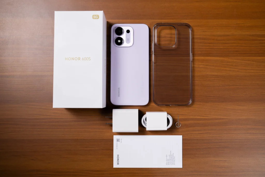 An unboxing flat lay of the purple HONOR 600S 5G showing the retail box, phone, clear protective case, charging adapter, USB cable, SIM ejector tool, and user documentation on a wooden desk.