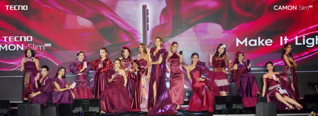 A wide-angle group photo on stage featuring 13 models posing in various shades of burgundy and maroon formal attire, each holding a smartphone. The background shows a large vibrant pink and red screen display with "TECNO CAMON Slim 5G" branding and the slogan "Make It Lighter".