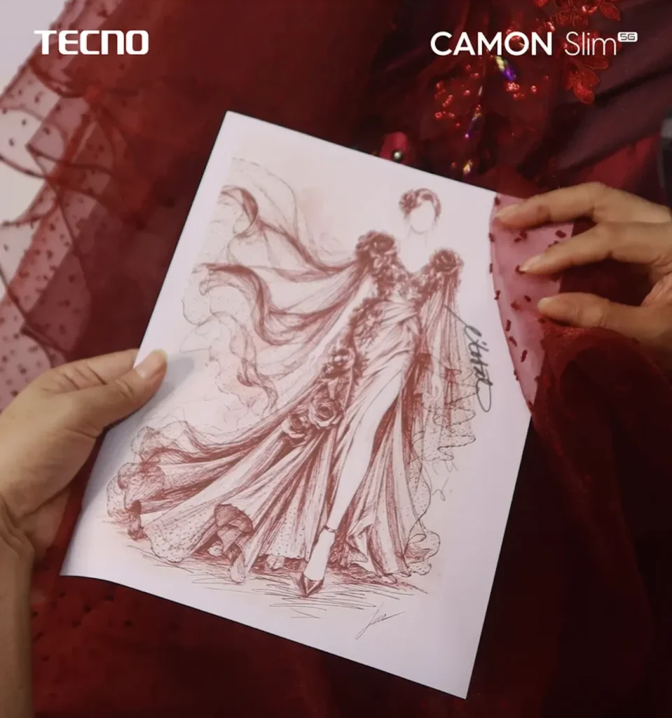 A pair of hands holds a detailed fashion illustration sketch of an elegant burgundy gown featuring floral shoulder accents, a thigh-high slit, and a flowing cape-like overlay. The sheet rests against a rich red, textured fabric with subtle embellishments. TECNO and CAMON Slim 5G branding appear in the top corners.